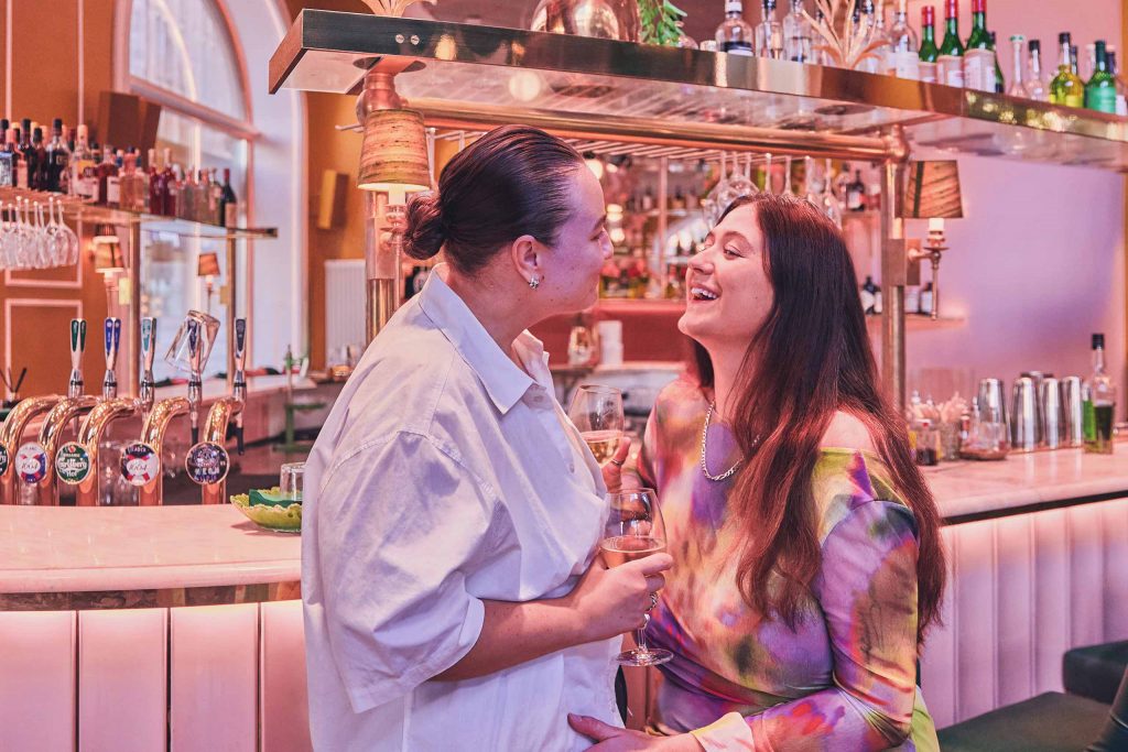 A lesbian couple staring into each other's eyes at a pink bar.