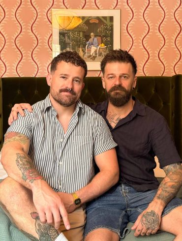 A gay couple sat on a bed in a hotel suite.