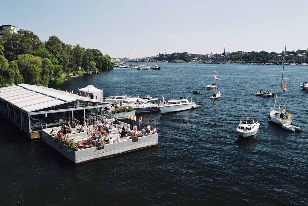 An aerial view of a floating waterfront restaurant on a sunny day, with people dining on the terrace as sailboats and motorboats decorated with Pride flags drift across a wide blue bay.