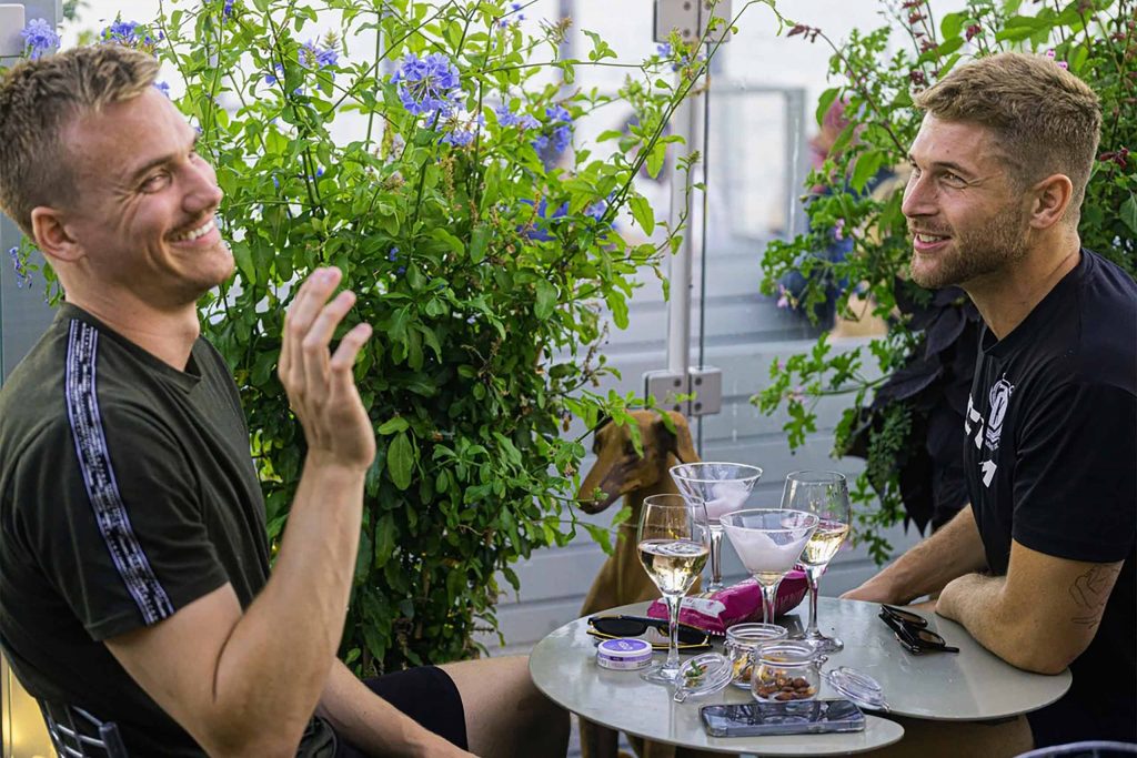 Two people sit at an outdoor table surrounded by lush plants, smiling and chatting over drinks on the restaurant's waterside terrace.