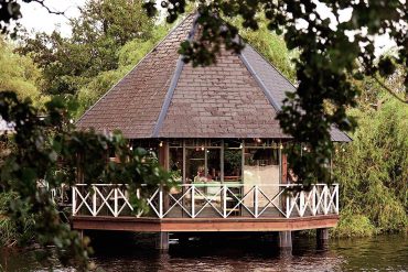 A cosy wooden pavilion with a steep, pyramid-shaped roof stands above the water, framed by leafy trees and reflected in the calm surface below.