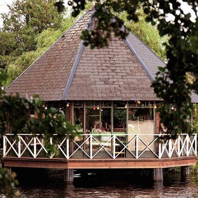A cosy wooden pavilion with a steep, pyramid-shaped roof stands above the water, framed by leafy trees and reflected in the calm surface below.
