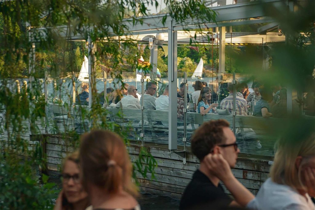 People enjoy food and drinks on a lively waterside terrace, viewed through leafy trees, with sailboats gliding across the water in the background.