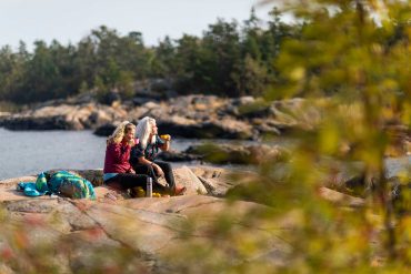 Two women in hiking gear sit on a rock looking out to sea, with a cup of tea taking in the majesty of the Stockholm Archipelago