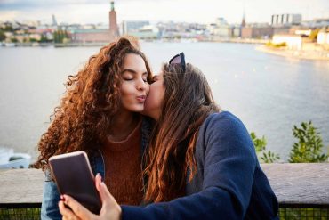 Two women with long brown hair kiss each other while taking a selfie with the backdrop of the city of Stockholm