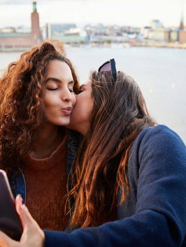 Two women with long brown hair kiss each other while taking a selfie with the backdrop of the city of Stockholm