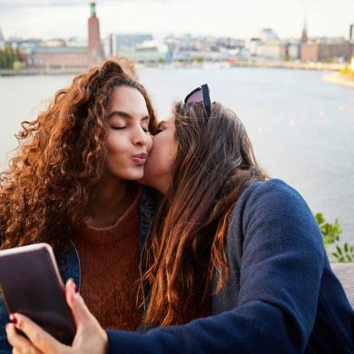 Two women with long brown hair kiss each other while taking a selfie with the backdrop of the city of Stockholm