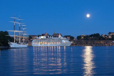 Cruise ship in Stockholm harbour, next to large sail boat in the late evening with a full moon in the sky reflected in the water