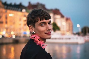 Queer person with a pink feather earring smiles to camera with the waterfront of Stockholm behind them