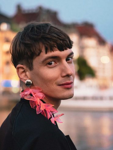 Queer person with a pink feather earring smiles to camera with the waterfront of Stockholm behind them