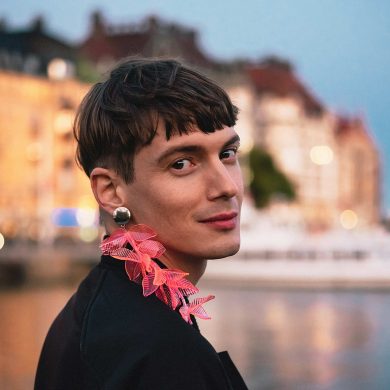 Queer person with a pink feather earring smiles to camera with the waterfront of Stockholm behind them