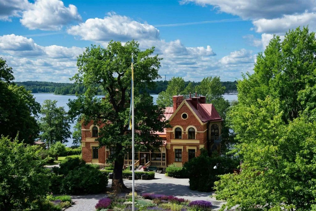 Exterior of Hotel J surrounded by greenery. A waterway is behind the hotel. Hotel J is ones of the best places to enjoy summer in Stockholm.