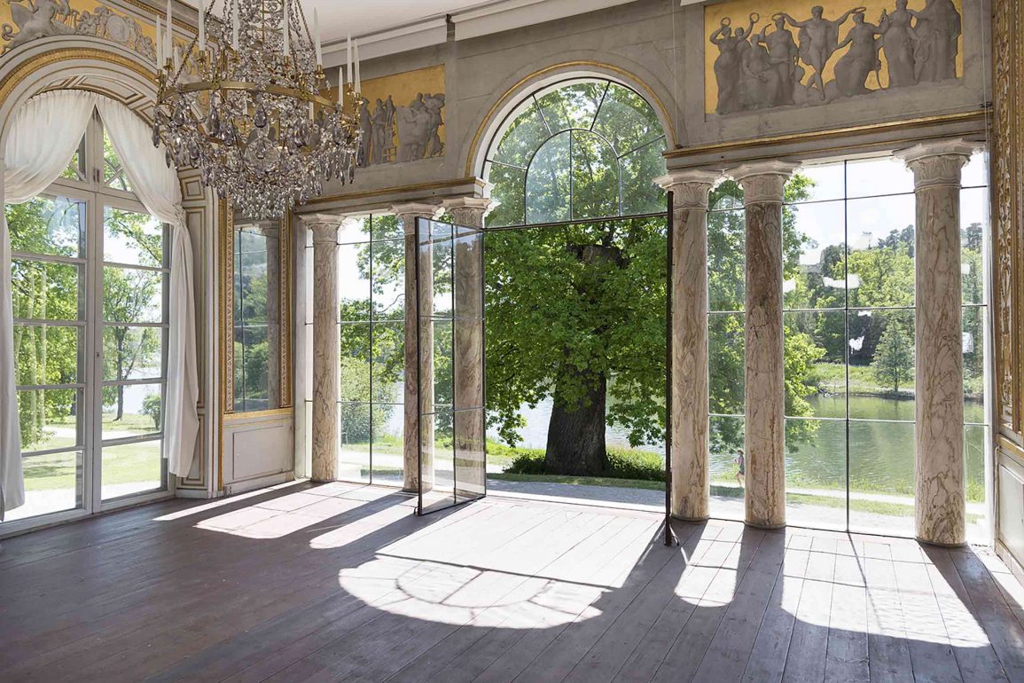 Sunlight shines into the Gustav III's Pavilion in Haga Park, outside a tree and a lake, inside an ornate white marble pavilon with chandelier
