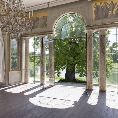 Sunlight shines into the Gustav III's Pavilion in Haga Park, outside a tree and a lake, inside an ornate white marble pavilon with chandelier