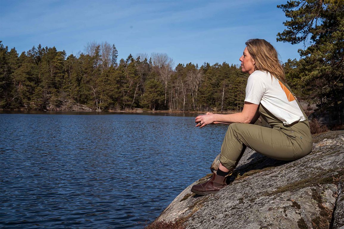 A woman sits on a rock overlooking a lake. The lake is surrounded by a forest.